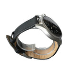 A stainless steel wristwatch with a black dial and black leather strap, displayed on a clear acrylic stand against a textured gray background.